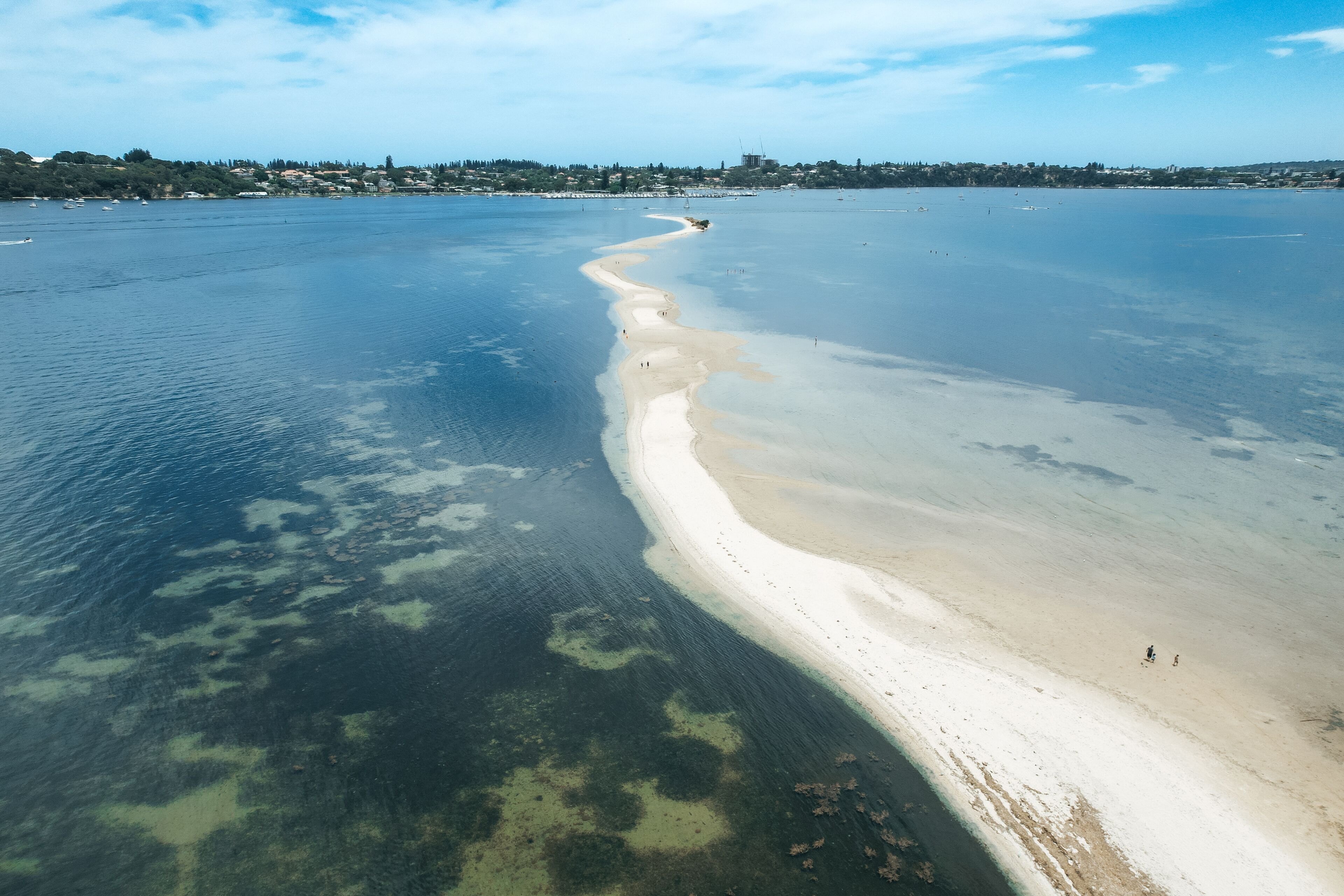 An aerial view of a beach with clear water, a sandy shoreline, and a distant cityscape.