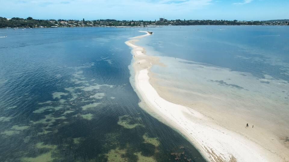 An aerial view of a beach with clear water, a sandy shoreline, and a distant cityscape.