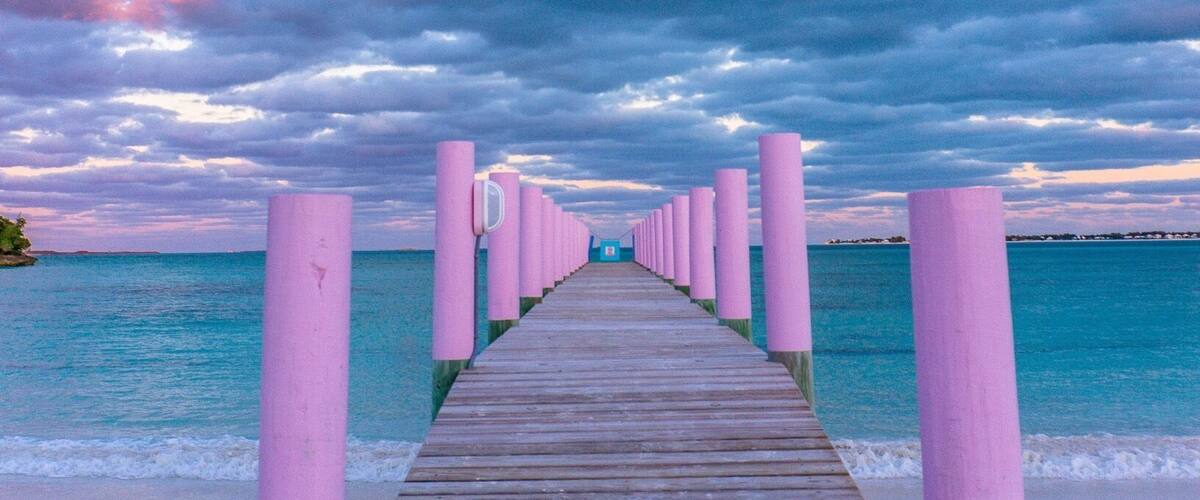 A wooden pier leading to a beach with pink posts, under a cloudy sky.