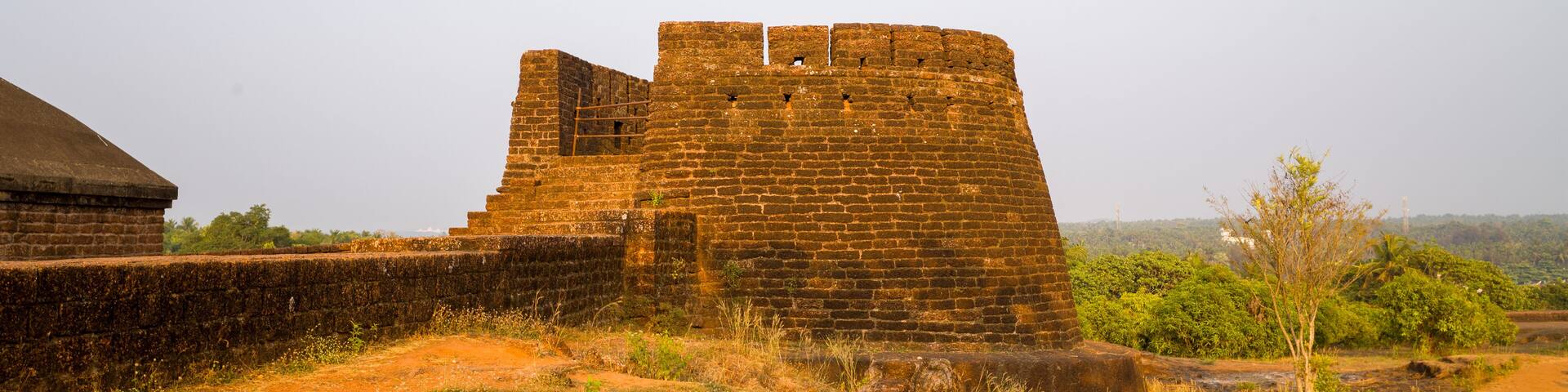 A robust stone bastion rises above sunlit fort walls at Bekal Fort, India, surrounded by dry earth and sparse greenery. Warm evening light casts a golden glow on the textured masonry and open