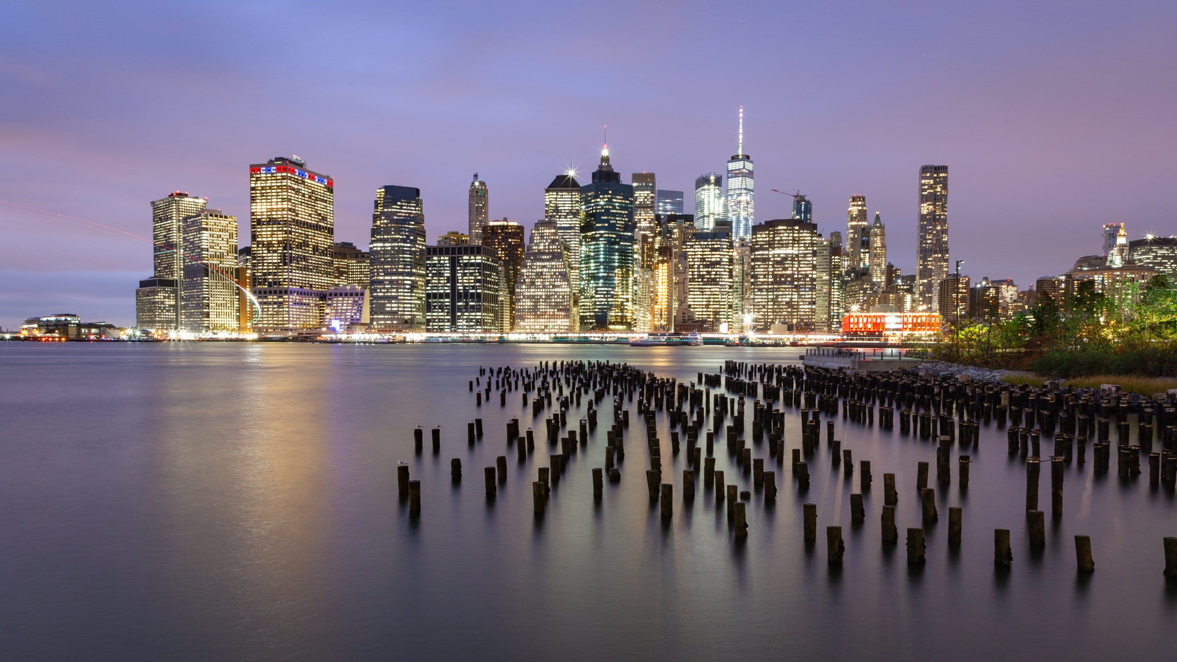 A city skyline at dusk with illuminated buildings and a calm water body with wooden posts.
