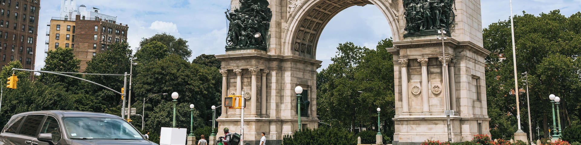 A large stone arch monument with statues on top, surrounded by trees and buildings.