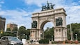 Un gran monumento de piedra en forma de arco con estatuas en la parte superior, rodeado de árboles y edificios.