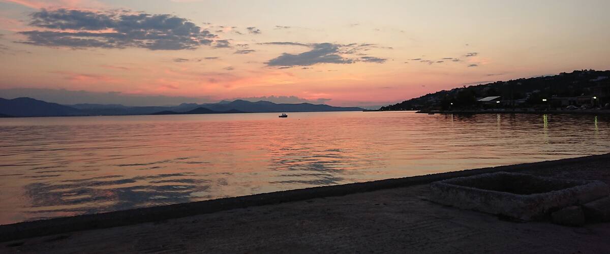 A sunset over a calm lake with a boat in the distance and a hilly shoreline with buildings.
