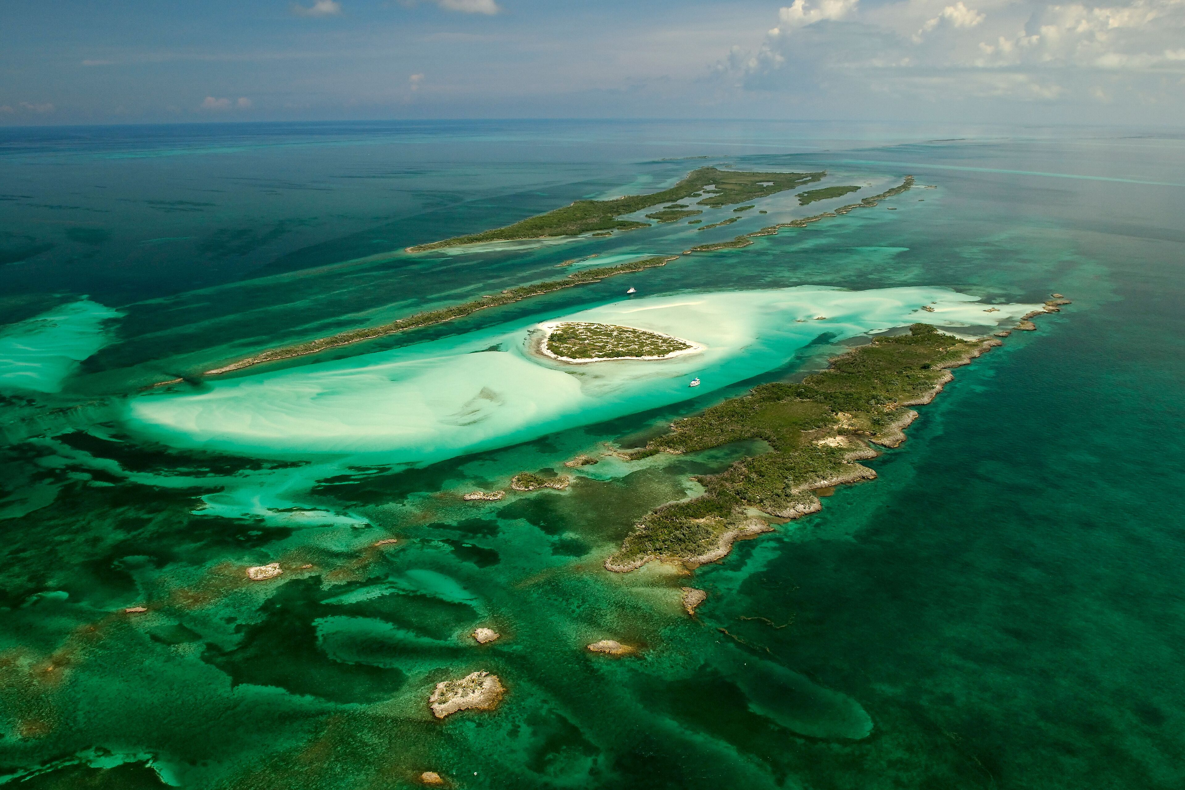 An aerial view of a coral reef with clear turquoise waters, a small island with a white sandy beach, and a boat in the distance.