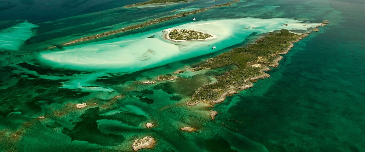 An aerial view of a coral reef with clear turquoise waters, a small island with a white sandy beach, and a boat in the distance.