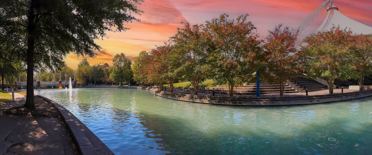 A riverside park with trees, a bridge, and a modern structure.