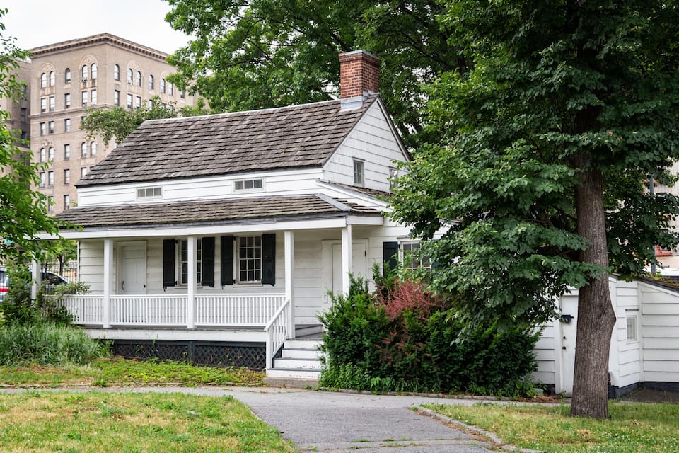 A white colonial-style house with a porch, surrounded by trees and a brick building in the background.