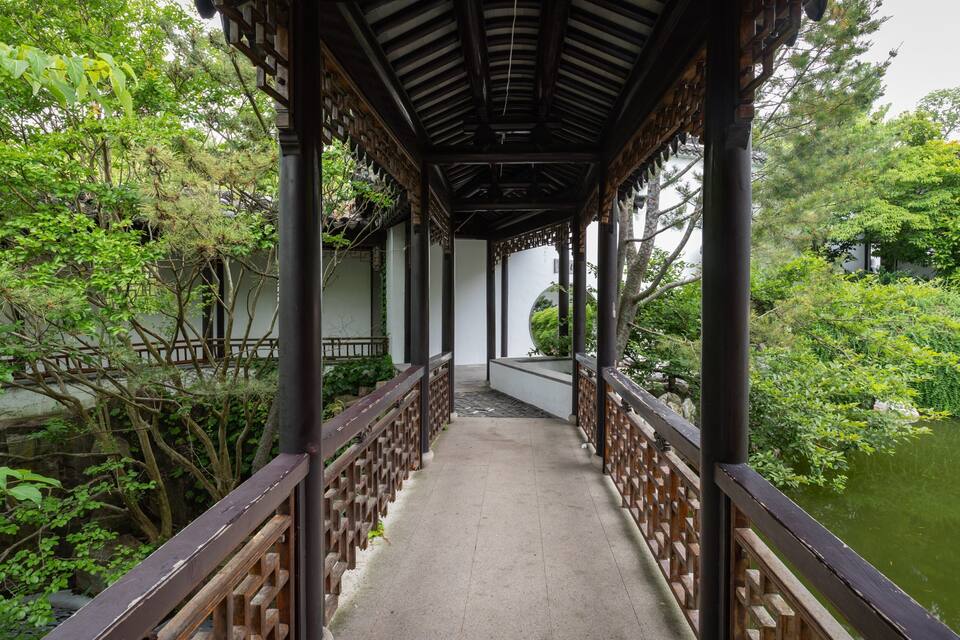 A wooden bridge over a pond in a garden with lush greenery and a traditional pavilion.