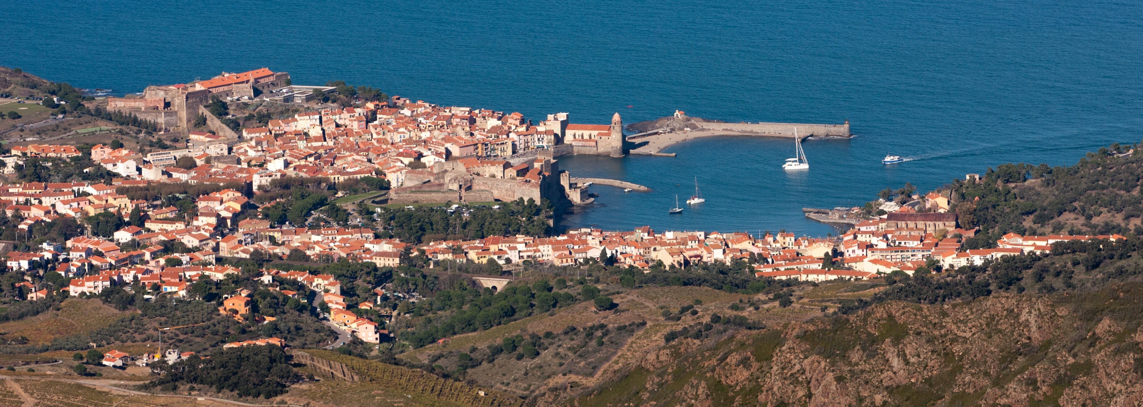 A coastal town with a historic fort, a harbor with boats, and a dense cluster of red-roofed buildings.