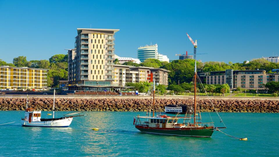 A harbor with two boats, a city skyline with modern buildings, and a rocky breakwater.