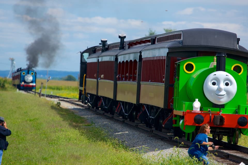 Un tren modelo con la parte delantera verde y un vagón central rojo y beige, sobre vías con una persona fotografiando y otra cerca de las vías.