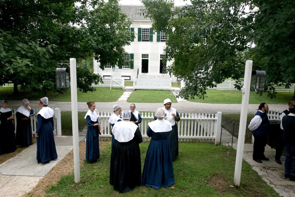A group of people in traditional attire standing in a grassy area with a white building in the background.