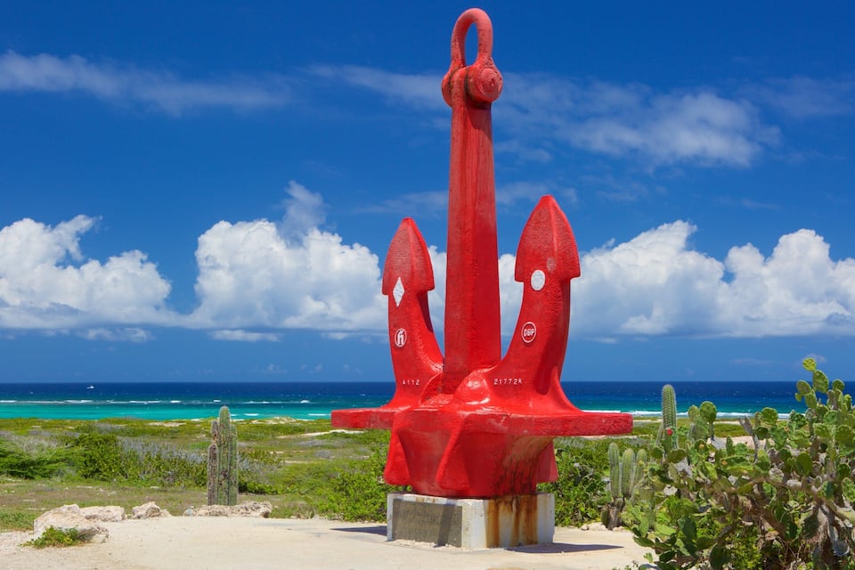 Una escultura de ancla roja con inscripciones, frente a un paisaje costero con cactus y un cielo azul despejado.