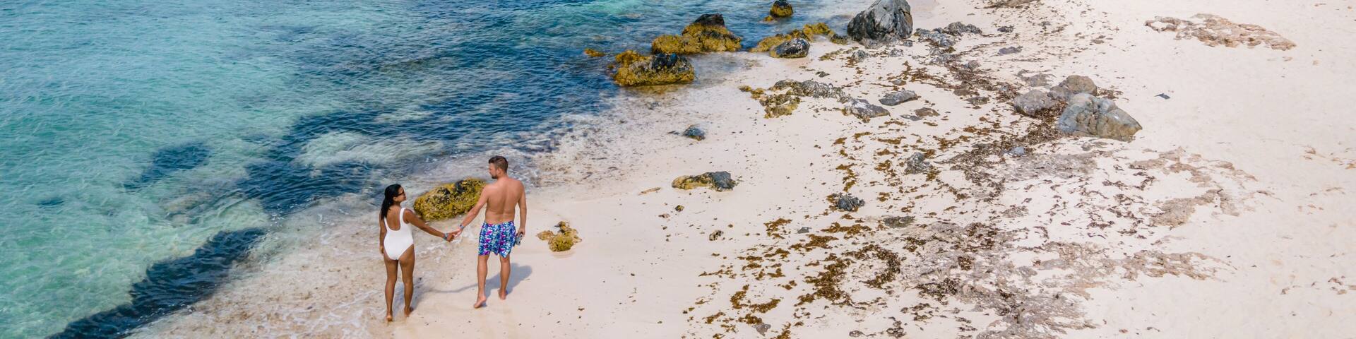 A beach with clear turquoise water, rocky shore, and boats in the distance.