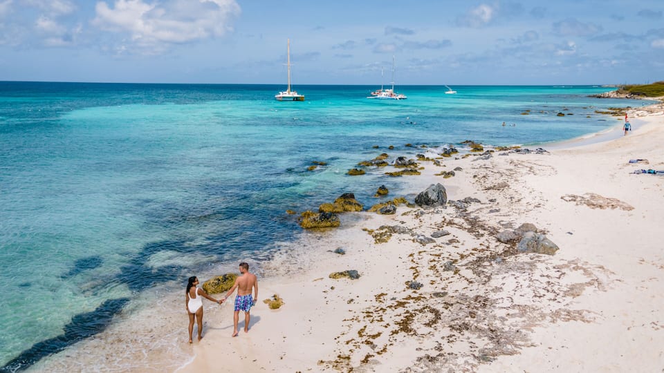 Une plage à l’eau turquoise et limpide, avec un rivage rocheux et des bateaux visibles au loin.