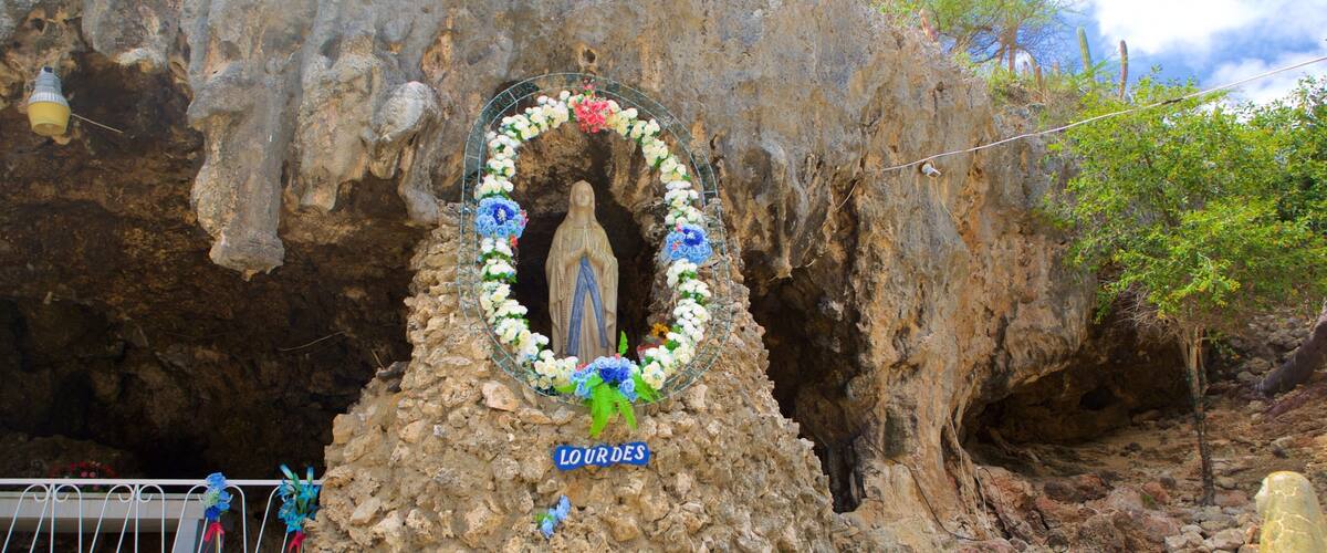 A statue of the Virgin Mary in Lourdes, surrounded by a floral wreath and a sign.
