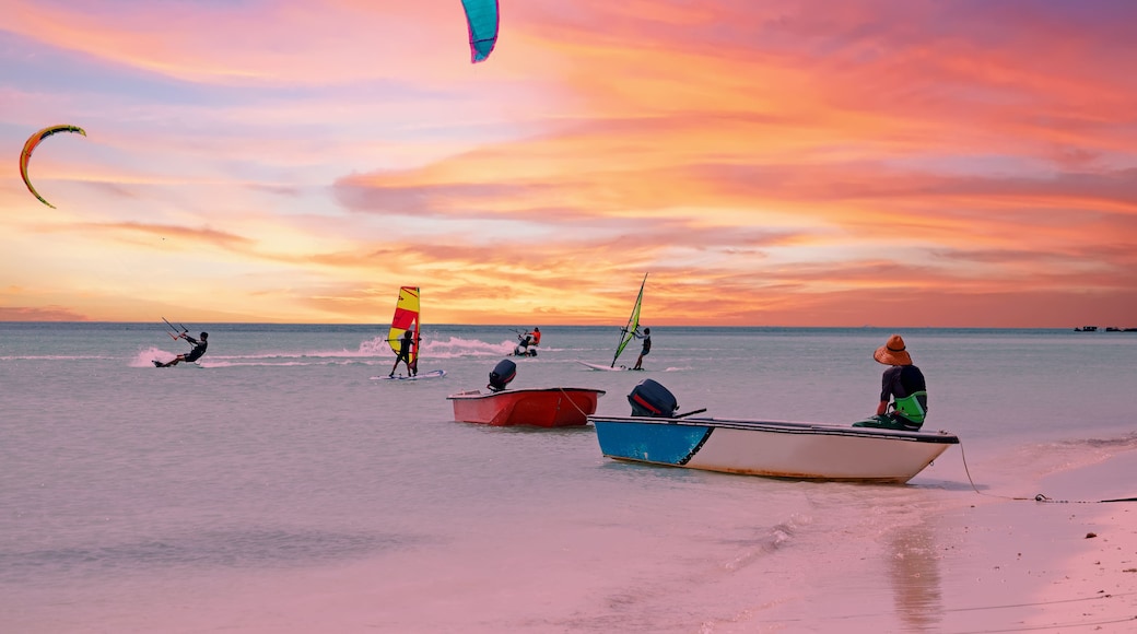 Uma praia com pessoas praticando windsurf e parapente ao pôr do sol.