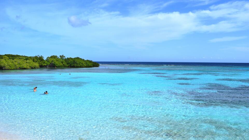 A clear turquoise sea with a sandy beach and a forested island in the distance.