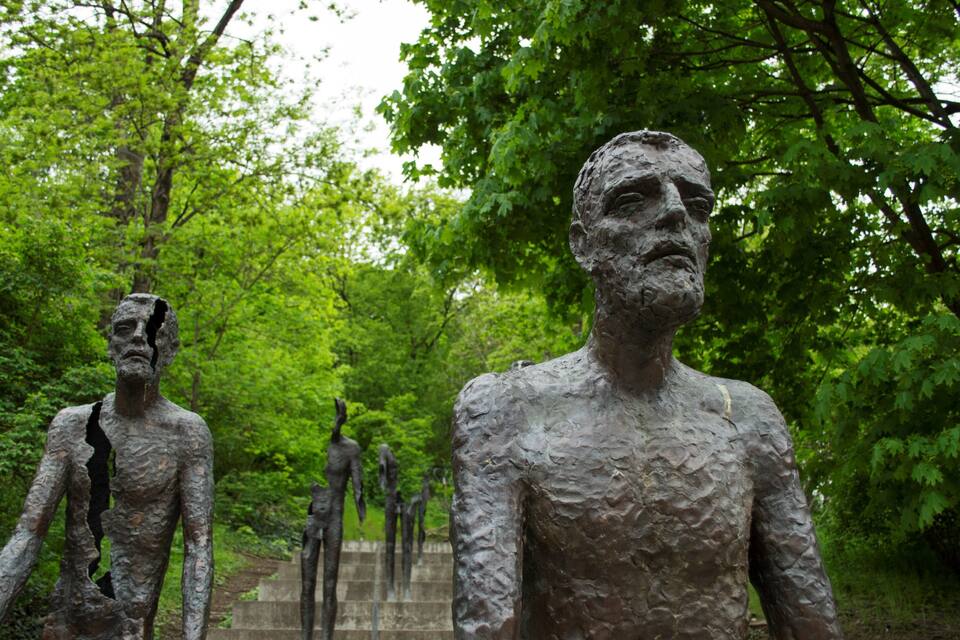 A sculpture garden with bronze statues of human figures set against a backdrop of lush green trees.