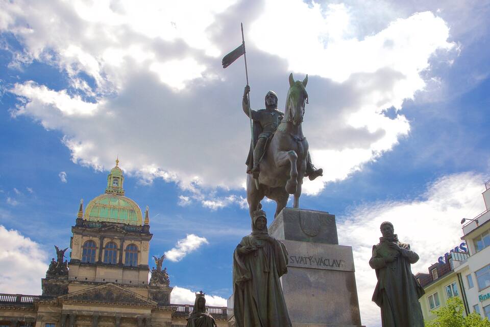Uma estátua de um homem a cavalo com uma bandeira, um edifício com cúpula e outra estátua em primeiro plano.