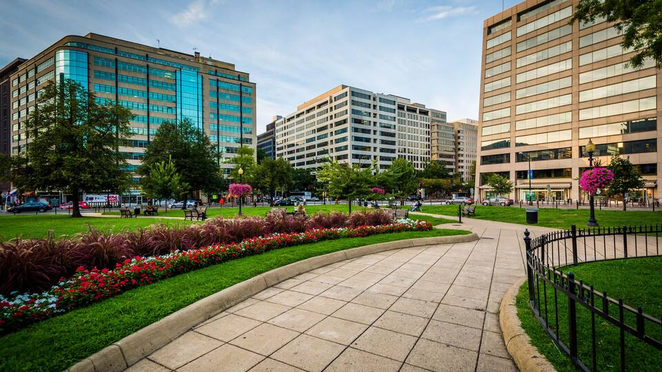 A city square with a curved pathway, surrounded by modern buildings, green lawns, and flowering plants.