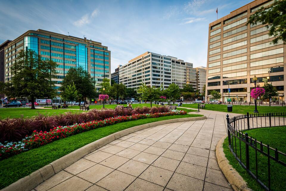 Uma praça urbana com um caminho curvo, rodeada por edifícios modernos, gramados verdes e plantas floridas.