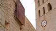 Ein verwittertes Schild mit der Aufschrift 'MUSTA ARQUITECTO' und einer Uhr an einem Steinhaus.