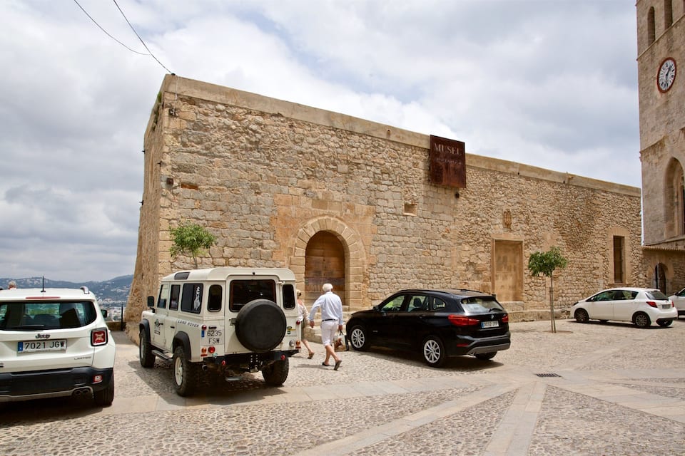 A stone building with a clock tower, parked cars, and a cobblestone street.
