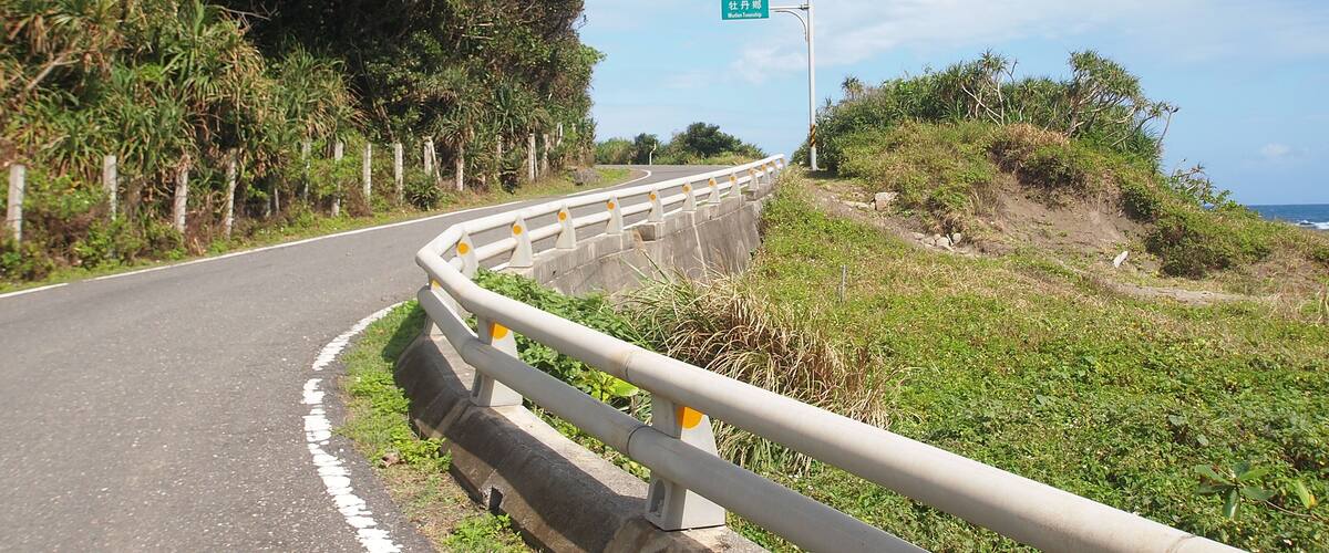 A winding road with a guardrail, surrounded by lush greenery and a clear blue sky.