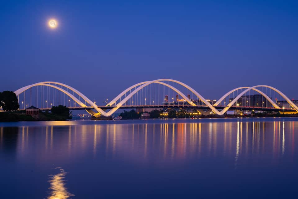 A bridge with a unique arch design, reflecting on a calm body of water under a night sky with a visible moon.