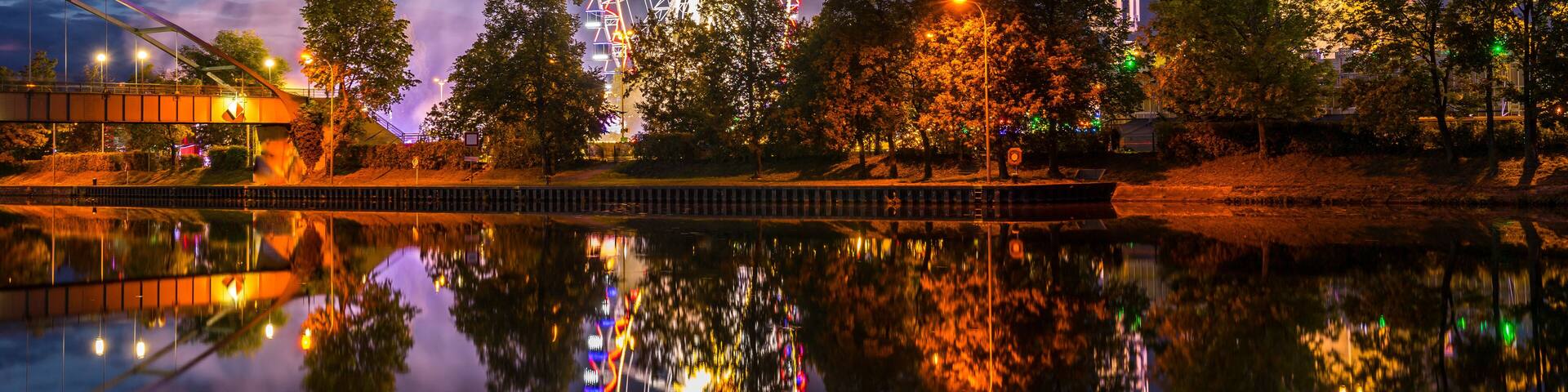 Une grande roue illuminée de mille feux se reflète dans l’eau calme d’un plan d’eau, entourée d’arbres et d’un pont.