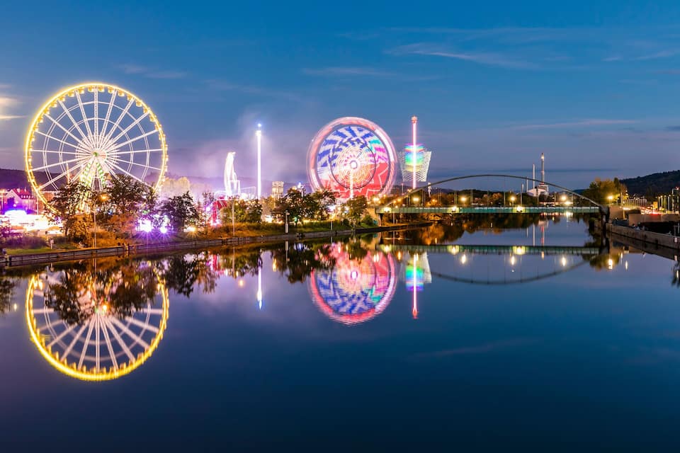 Een nachtzicht van een stad met twee grote reuzenraden, hun reflecties in kalm water, een brug en verlichte gebouwen.
