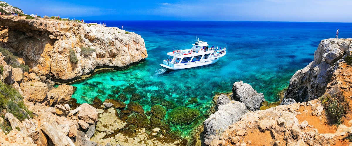 Cape Greco natural park boat trip in turquoise sea water Cyprus. Scenic panoramic view of tourist boat anchored between steep coastal rocks and stone arch lovers bridge in Ayia Napa.