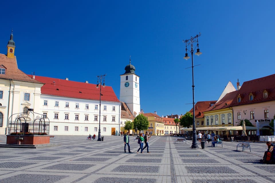 Uma praça histórica com calçada de paralelepípedos, uma torre do relógio imponente e arquitetura europeia tradicional.