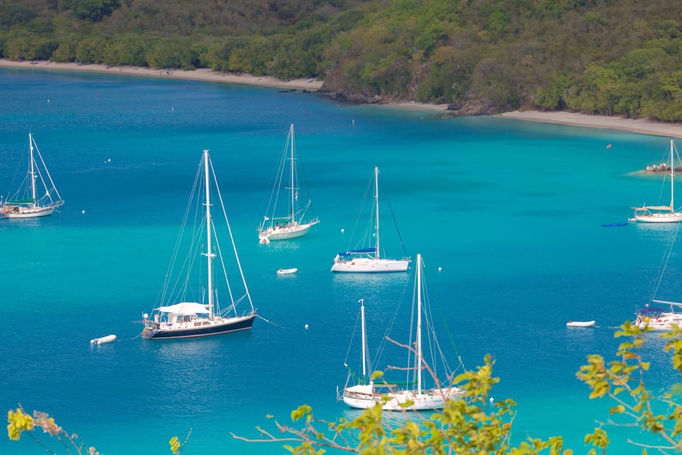 Een turquoise zee met verschillende zeilboten die voor anker liggen nabij een zandstrand, omringd door weelderige begroeiing.