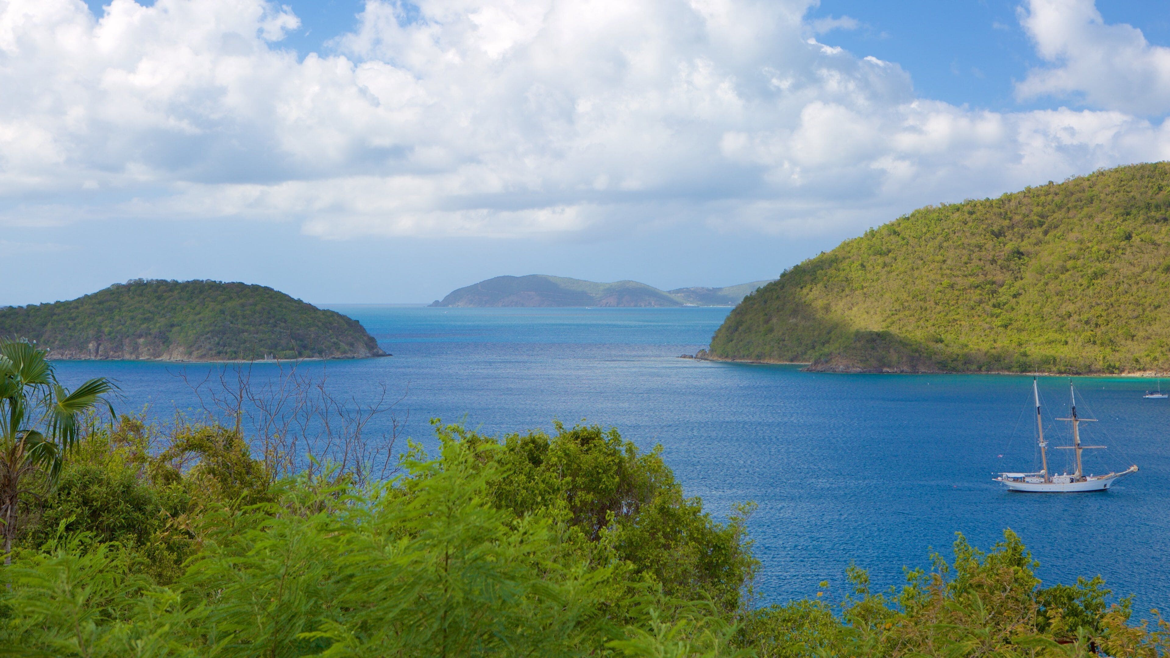 A coastal landscape with a clear blue sea, green hills, and a sailboat.