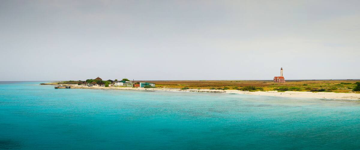 A coastal landscape with turquoise waters, a sandy beach, and a few buildings including a lighthouse.
