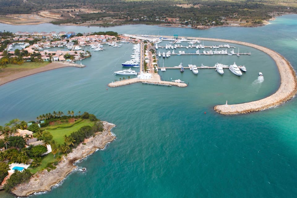 An aerial view of a marina with boats docked, surrounded by a coastal landscape with houses and greenery.