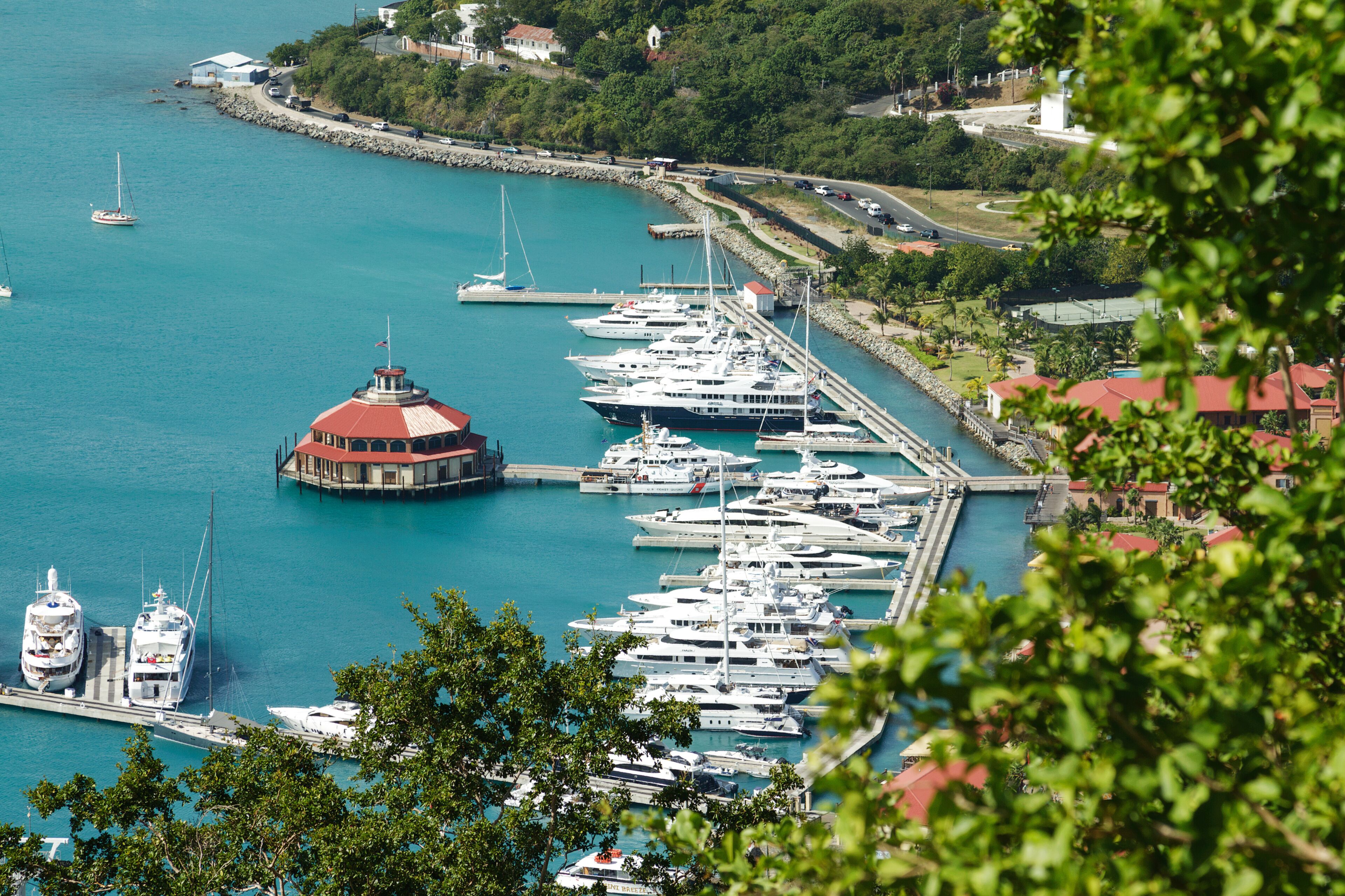 A marina with multiple yachts docked, a distinctive red-roofed building, and a clear turquoise sea.