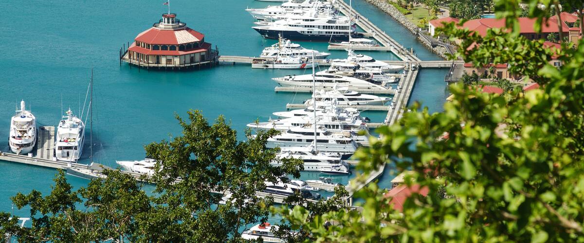 A marina with multiple yachts docked, a distinctive red-roofed building, and a clear turquoise sea.