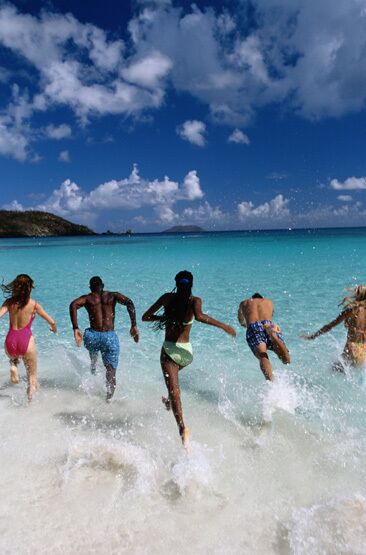 A group of people running into the sea on a beach with clear blue water and a clear sky.