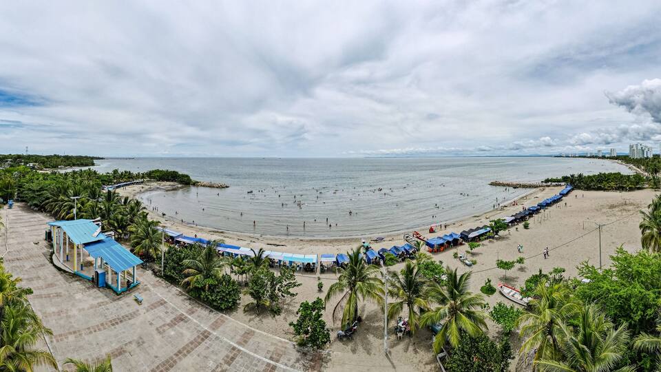 A beach with a curved shoreline, palm trees, and a row of blue-roofed structures along the water's edge.
