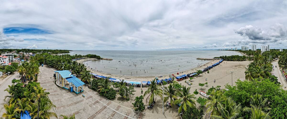 A beach with a curved shoreline, palm trees, and a row of blue-roofed structures along the water's edge.