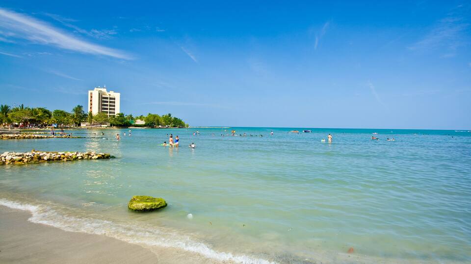A beach with clear turquoise water, a sandy shore, and a hotel in the background.