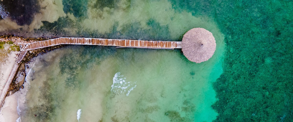 An aerial view of a coastal area with a wooden pier extending into turquoise waters, surrounded by a sandy beach and rocky outcrops.