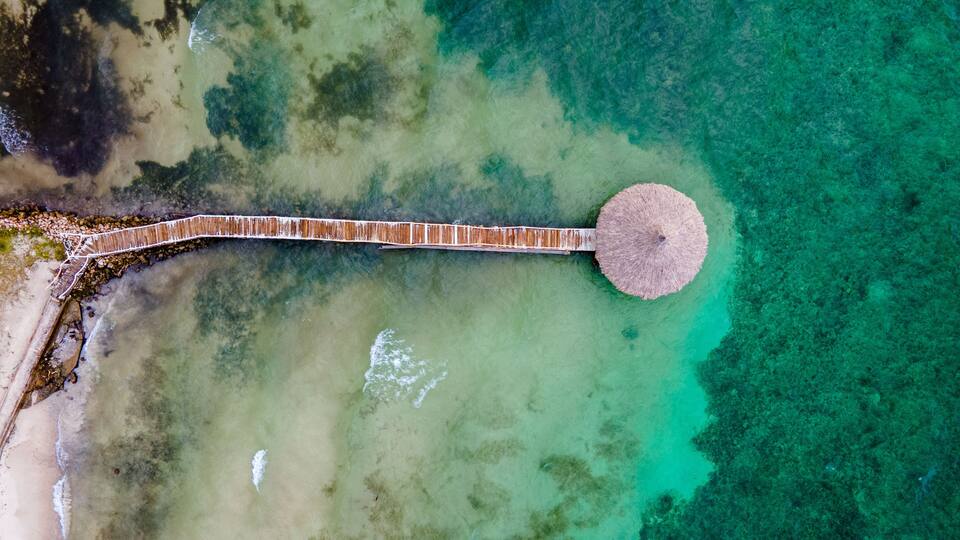 An aerial view of a coastal area with a wooden pier extending into turquoise waters, surrounded by a sandy beach and rocky outcrops.