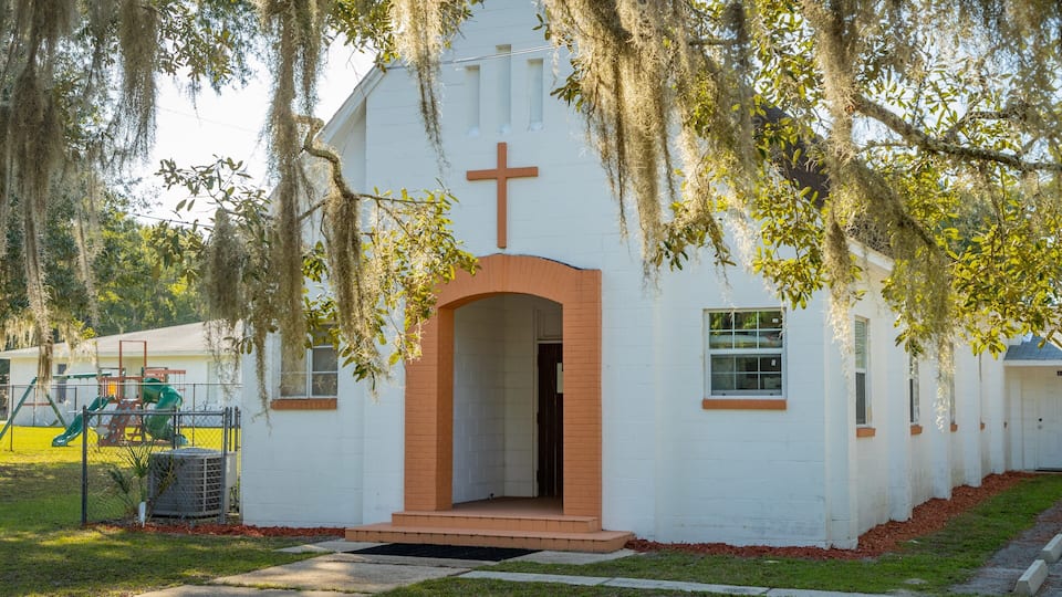 A white church with a cross, surrounded by trees and a playground.