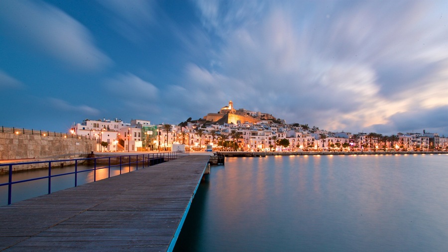 Un paisaje urbano costero al atardecer, con una fortaleza imponente en una colina, un muelle de madera y un frente marítimo tranquilo.