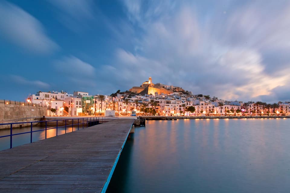 A coastal cityscape at dusk with a prominent hilltop fortress, a calm waterfront, and a wooden pier.